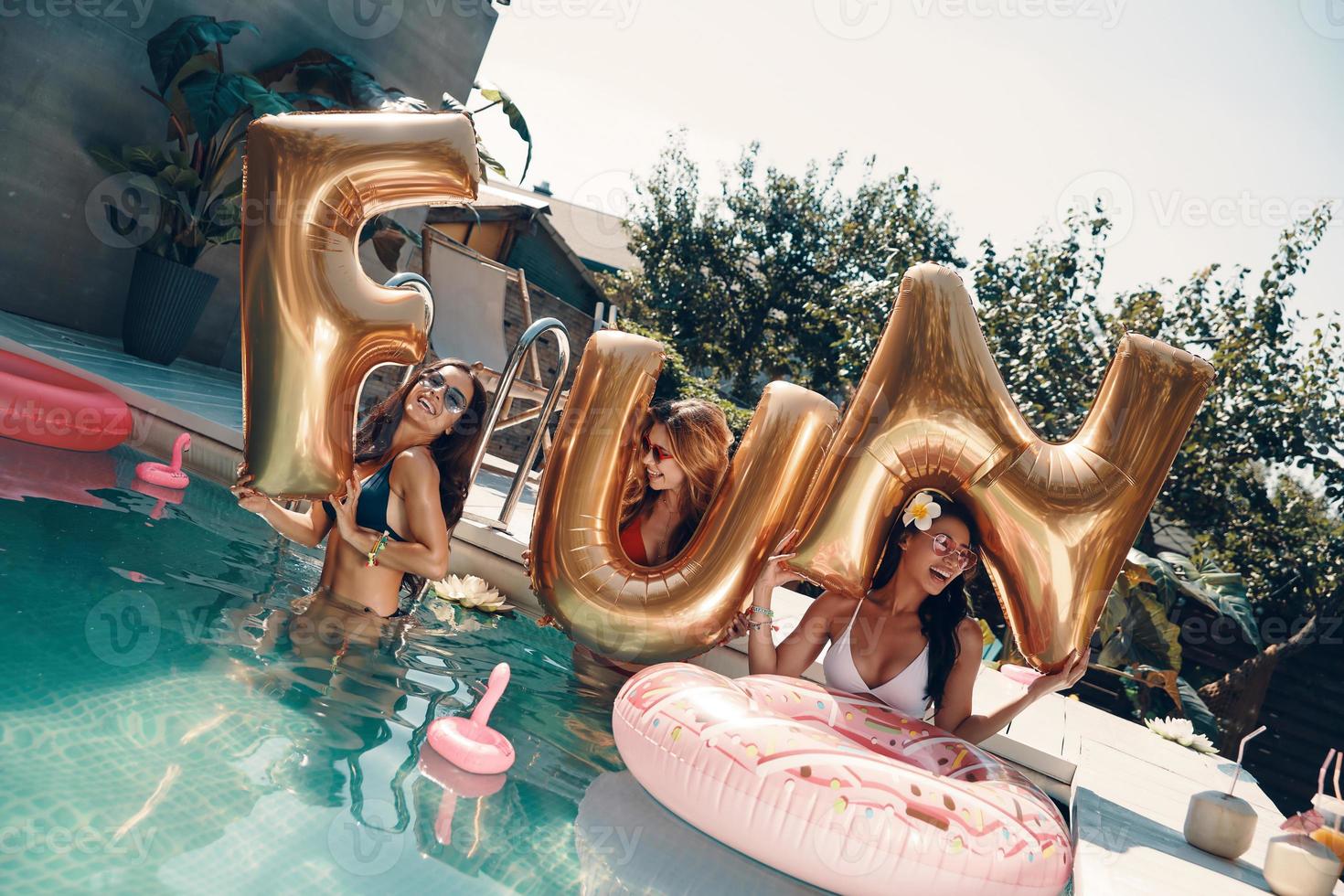 mulheres jovens atraentes em trajes de banho sorrindo e levantando balões em pé na piscina ao ar livre foto