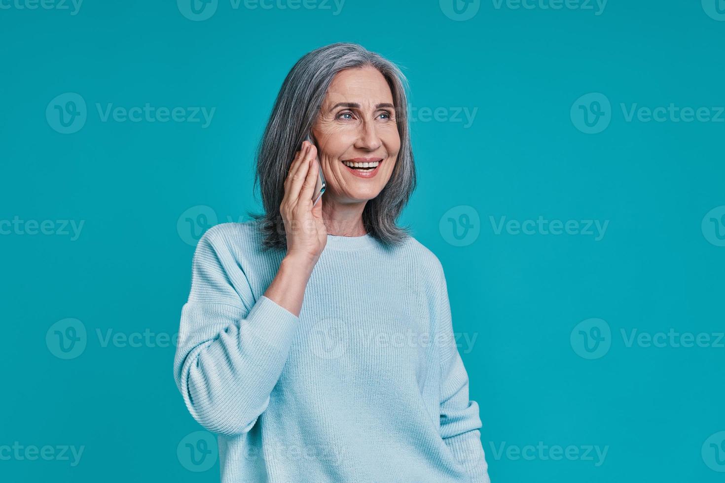 mulher bonita madura falando no telefone inteligente e sorrindo em pé contra um fundo azul foto