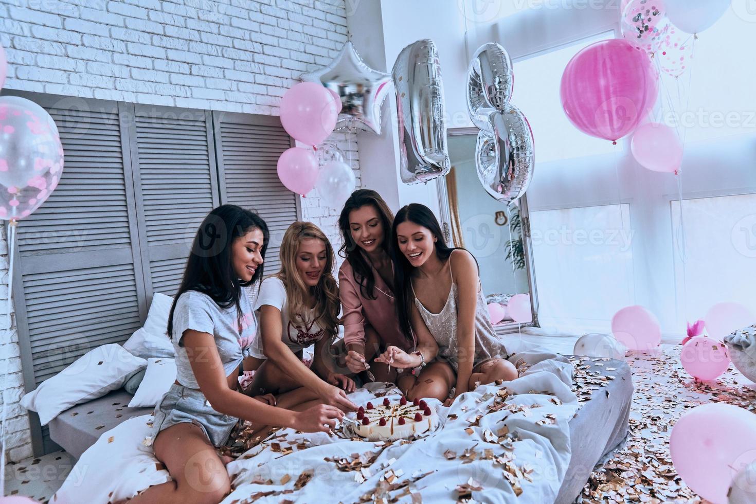hora do bolo. quatro belas jovens sorridentes de pijama comendo bolo enquanto faz uma festa do pijama no quarto com balões por todo o lado foto