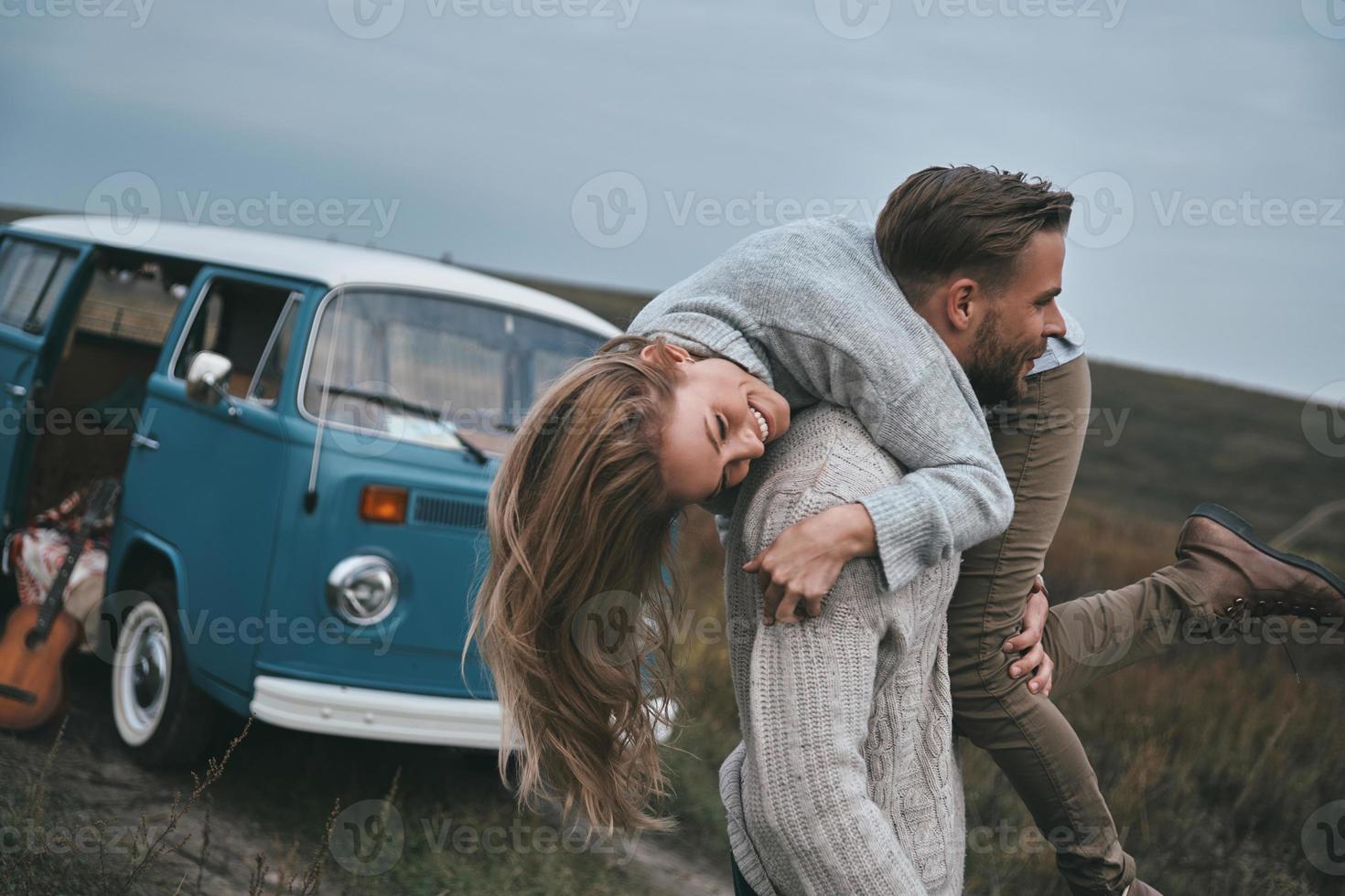 louco de amor. jovem bonito carregando sua namorada atraente nos ombros e sorrindo em pé perto da mini van estilo retrô azul foto