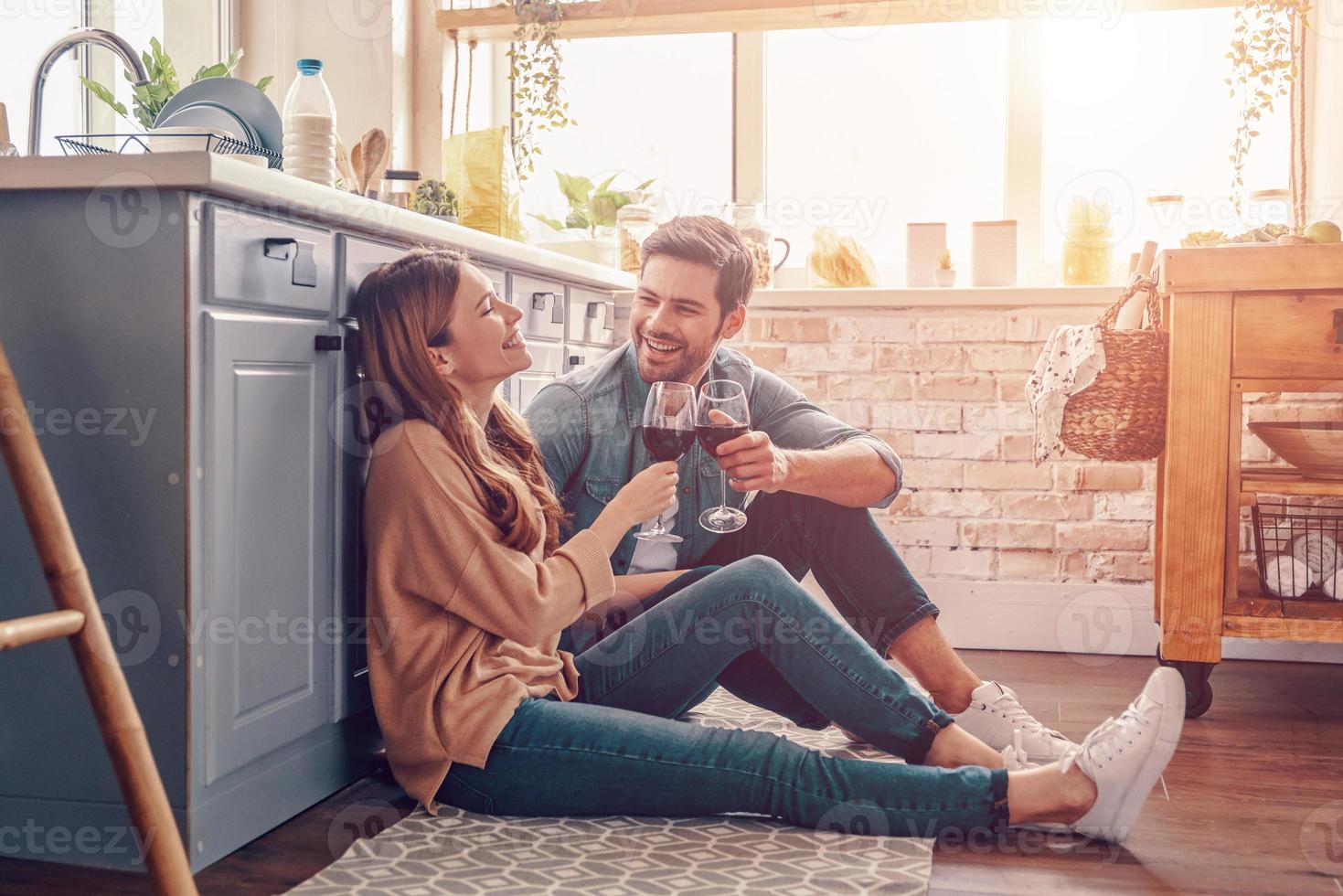 o amor está no ar. lindo casal jovem bebendo vinho enquanto está sentado no chão da cozinha em casa foto