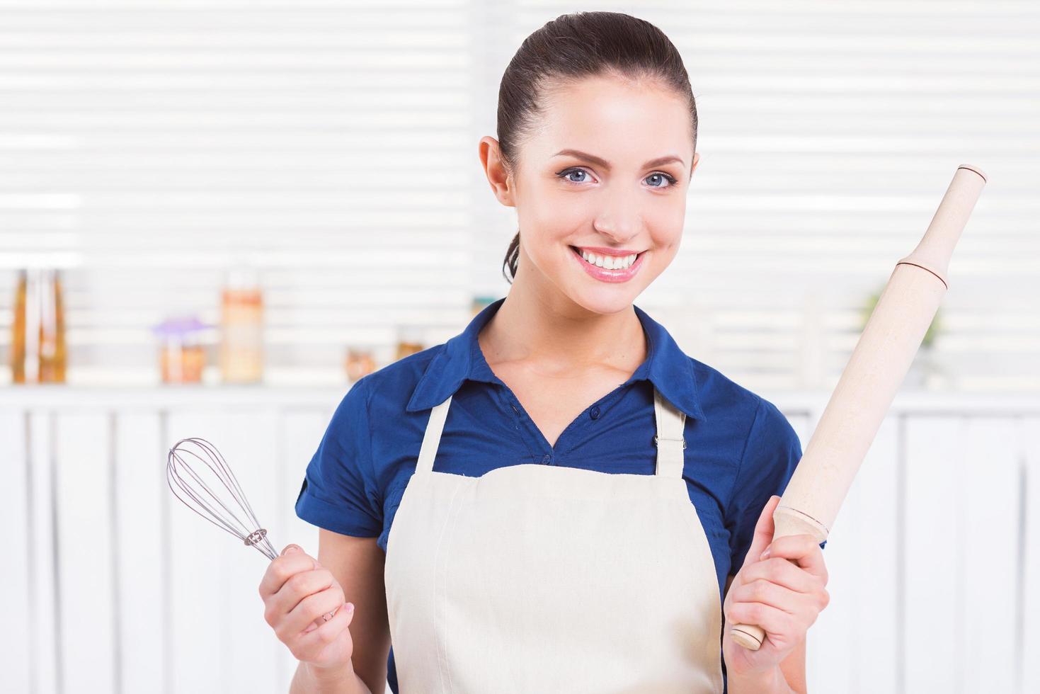 pronto para fazer pastel. mulher jovem e atraente no avental segurando o rolo e batedor de arame em pé em uma cozinha foto