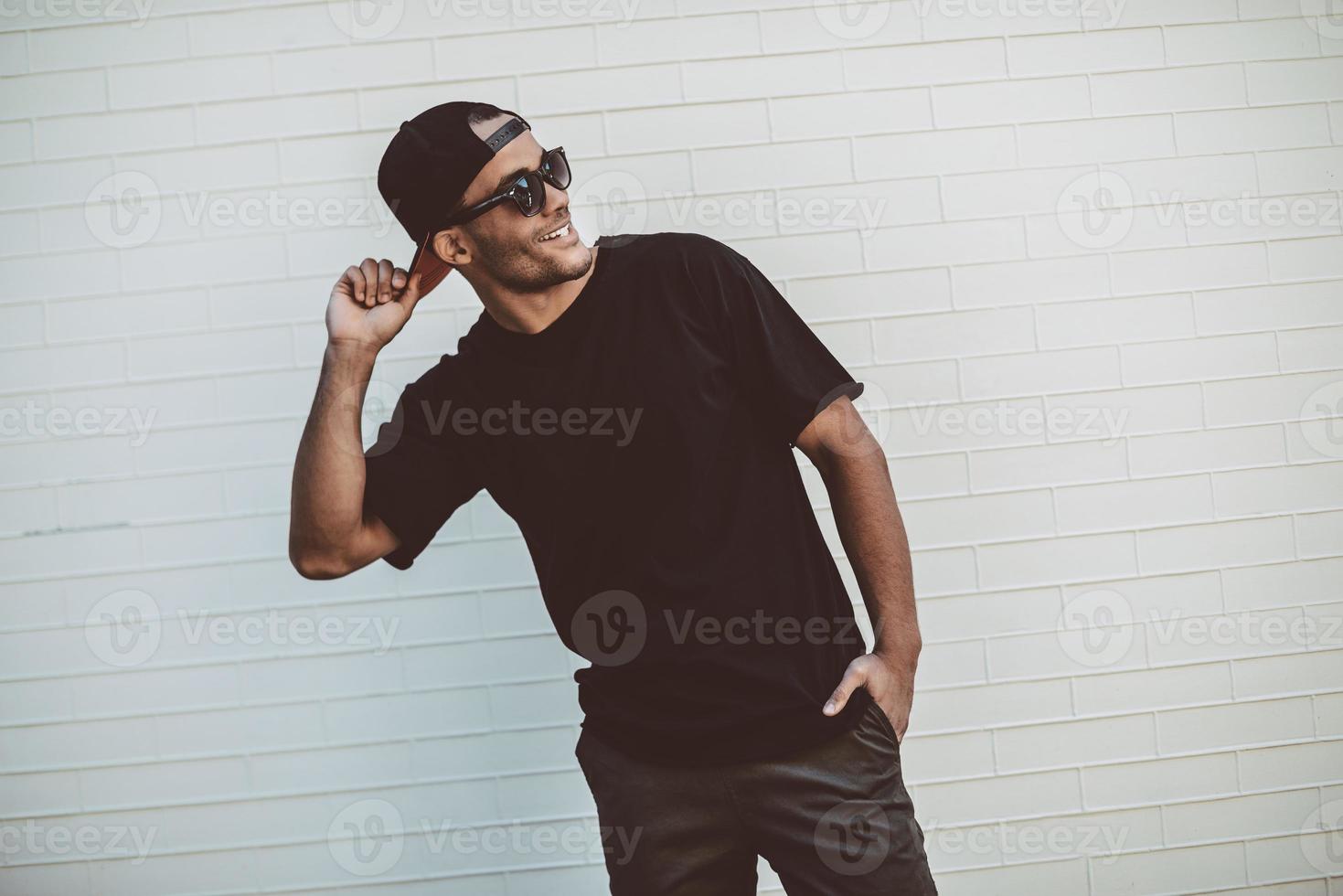 sempre de bom humor. jovem africano bonito em camiseta preta sorrindo e olhando para longe em frente à parede de tijolos ao ar livre foto