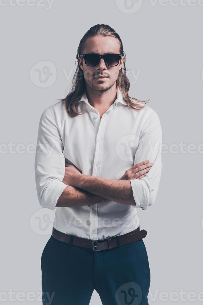 legal e bonito. jovem bonito na camisa branca e óculos de sol, olhando para a câmera e mantendo os braços cruzados em pé contra um fundo cinza foto