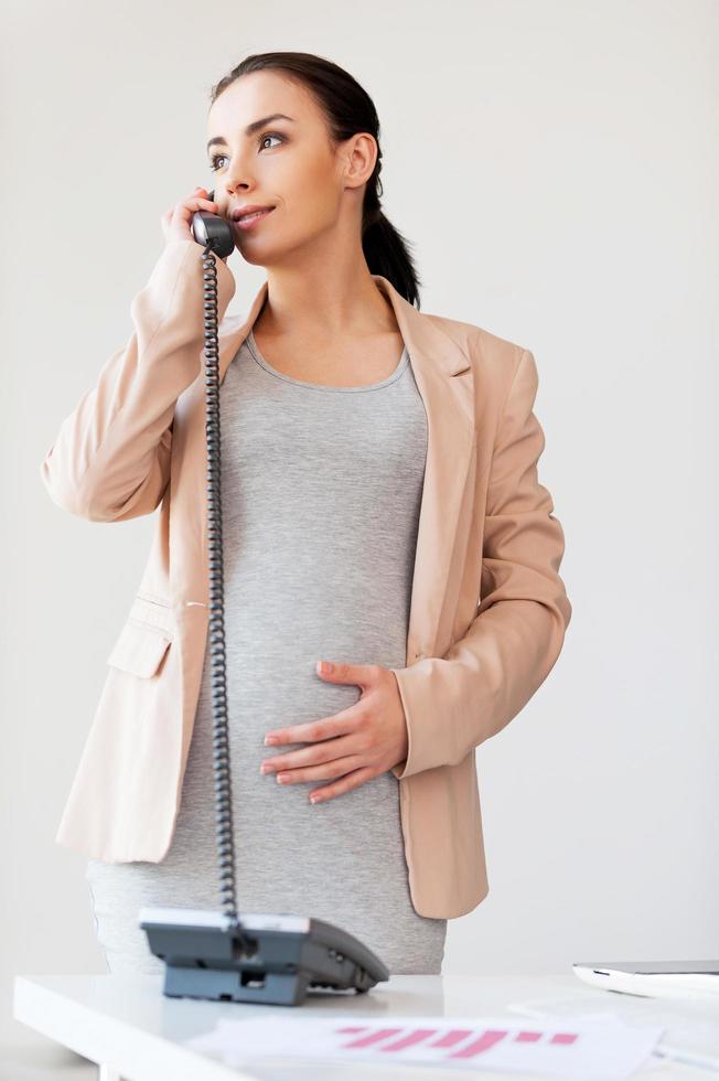 senhora de negócios grávida ao telefone. linda empresária grávida falando ao telefone e sorrindo em pé perto de seu local de trabalho no escritório foto