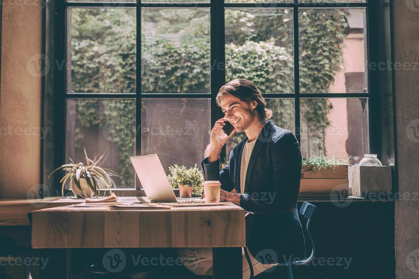 trabalhando do café. jovem alegre em roupas casuais inteligentes olhando para laptop e falando no celular enquanto está sentado perto da janela no escritório criativo ou café foto