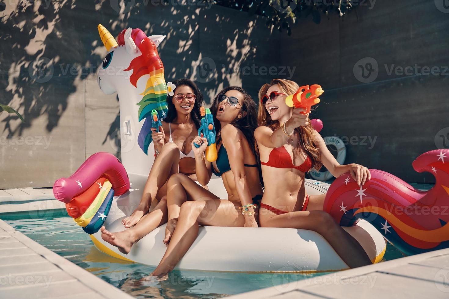 mulheres jovens atraentes em trajes de banho sorrindo e se divertindo com pistolas de água enquanto flutuava no unicórnio inflável na piscina ao ar livre foto
