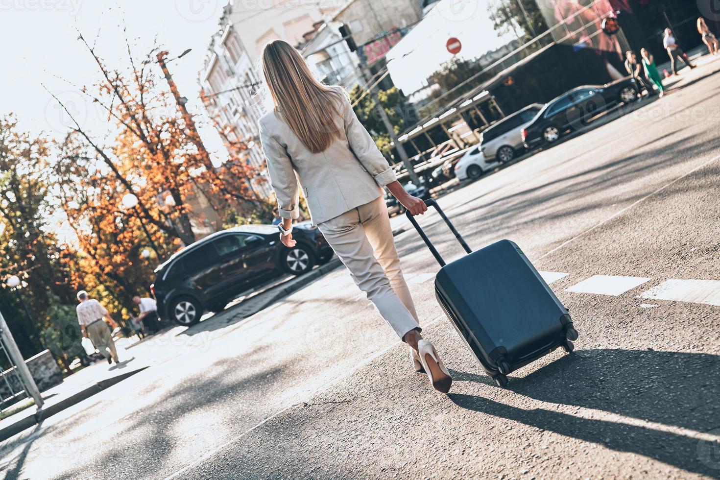 viagem de negócios. vista traseira de corpo inteiro de jovem de terno puxando bagagem enquanto caminhava ao ar livre foto