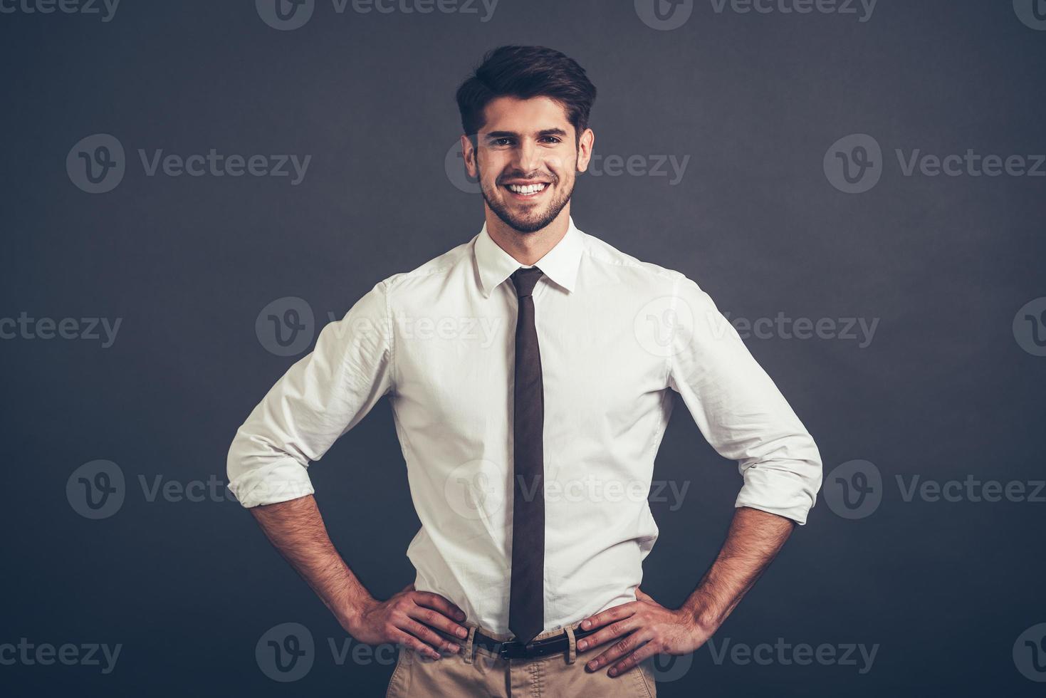 Melhor sorriso. confiante jovem bonito mantendo os braços akimbo e olhando para a câmera com sorriso em pé contra um fundo cinza foto