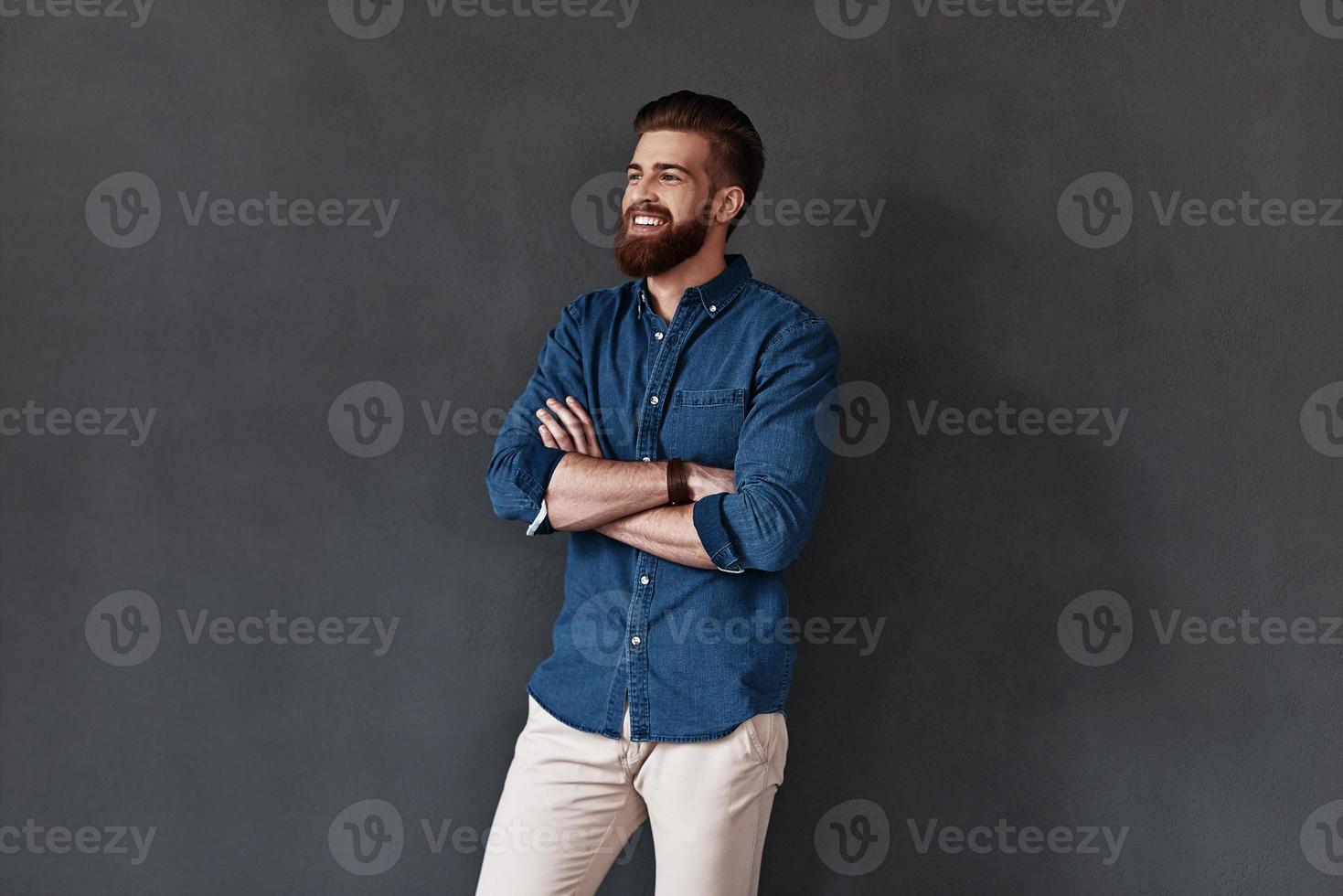 homem irresistível. braços de jovem bonito cruzados e desviar o olhar com sorriso em pé contra um fundo cinza foto