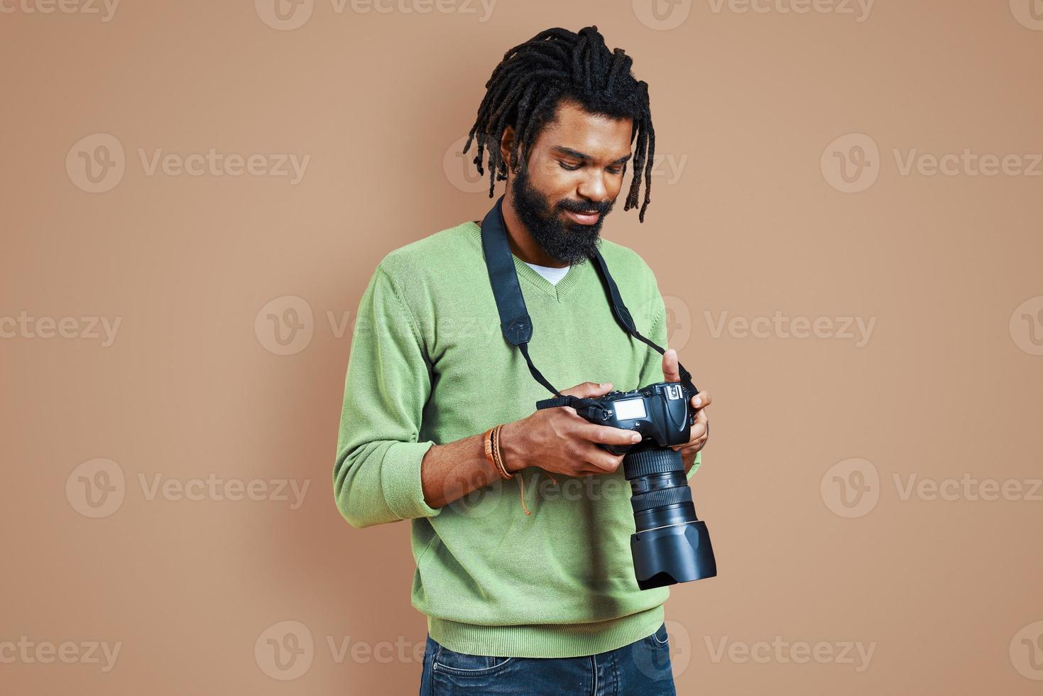 bonito jovem fotógrafo africano em roupas casuais usando câmera digital e sorrindo em pé contra um fundo marrom foto