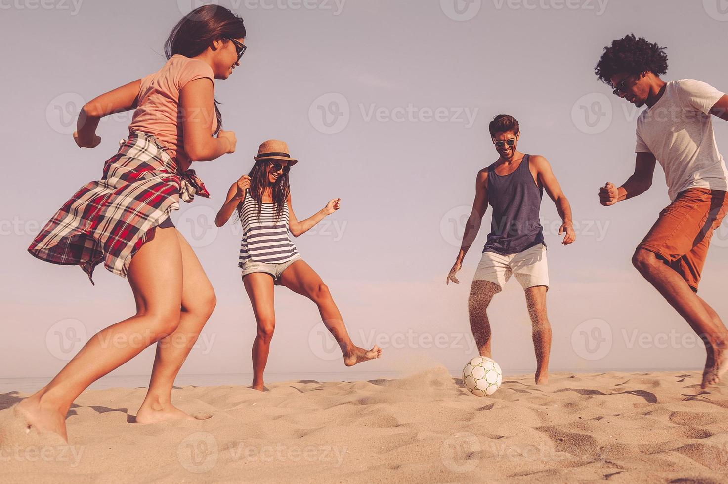 só me divertindo. grupo de jovens alegres brincando com bola de futebol na praia com mar ao fundo foto