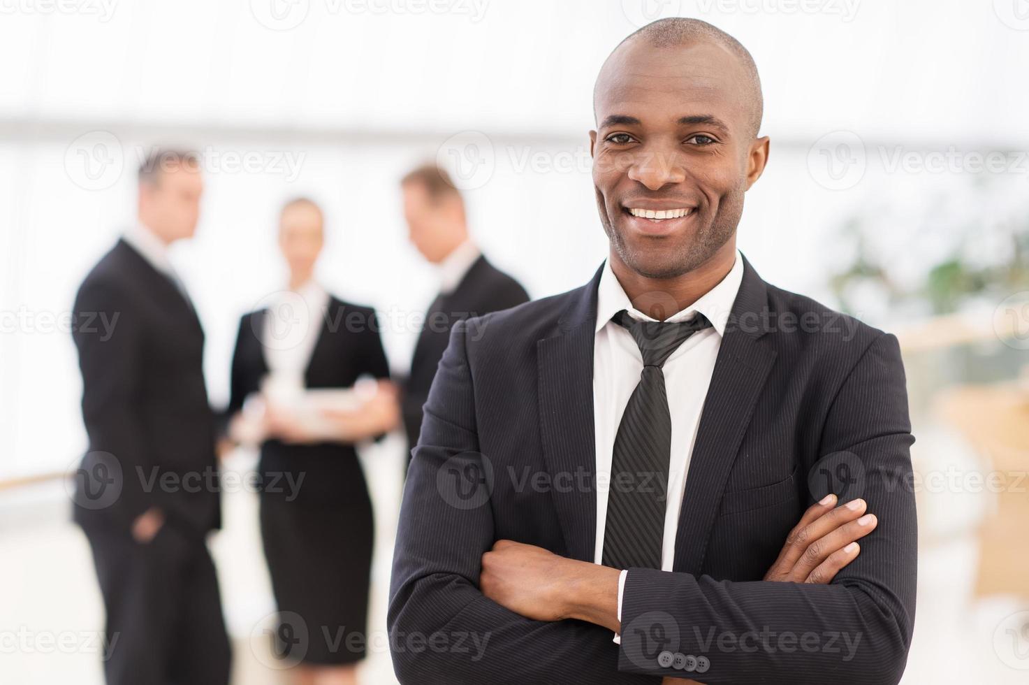 empresário confiante. alegre jovem africano em trajes formais, mantendo os braços cruzados e sorrindo enquanto seus colegas de pé no fundo foto