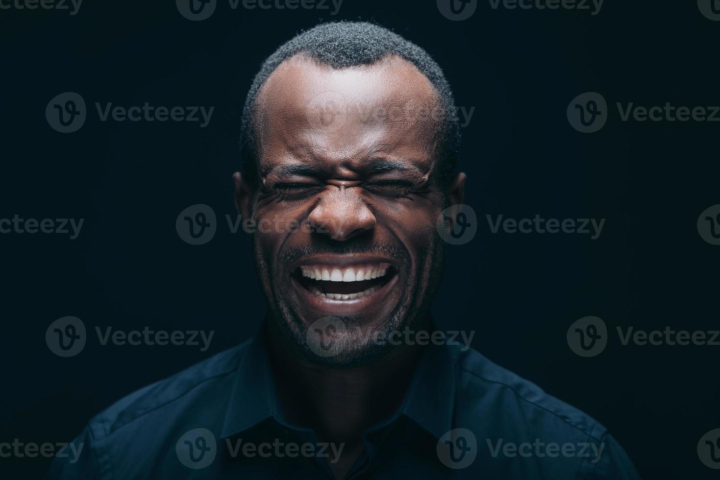 fazendo cara. retrato de jovem africano fazendo careta enquanto mantém os olhos fechados e está na frente de fundo preto foto