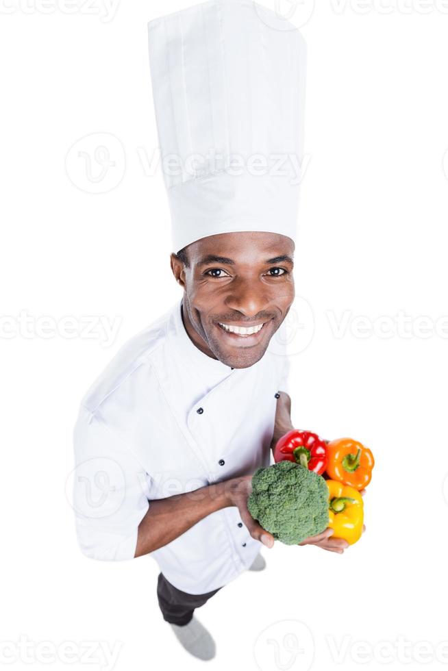 comida saudável está em suas mãos. vista superior do alegre jovem chef africano de uniforme branco segurando legumes coloridos em pé contra um fundo branco foto