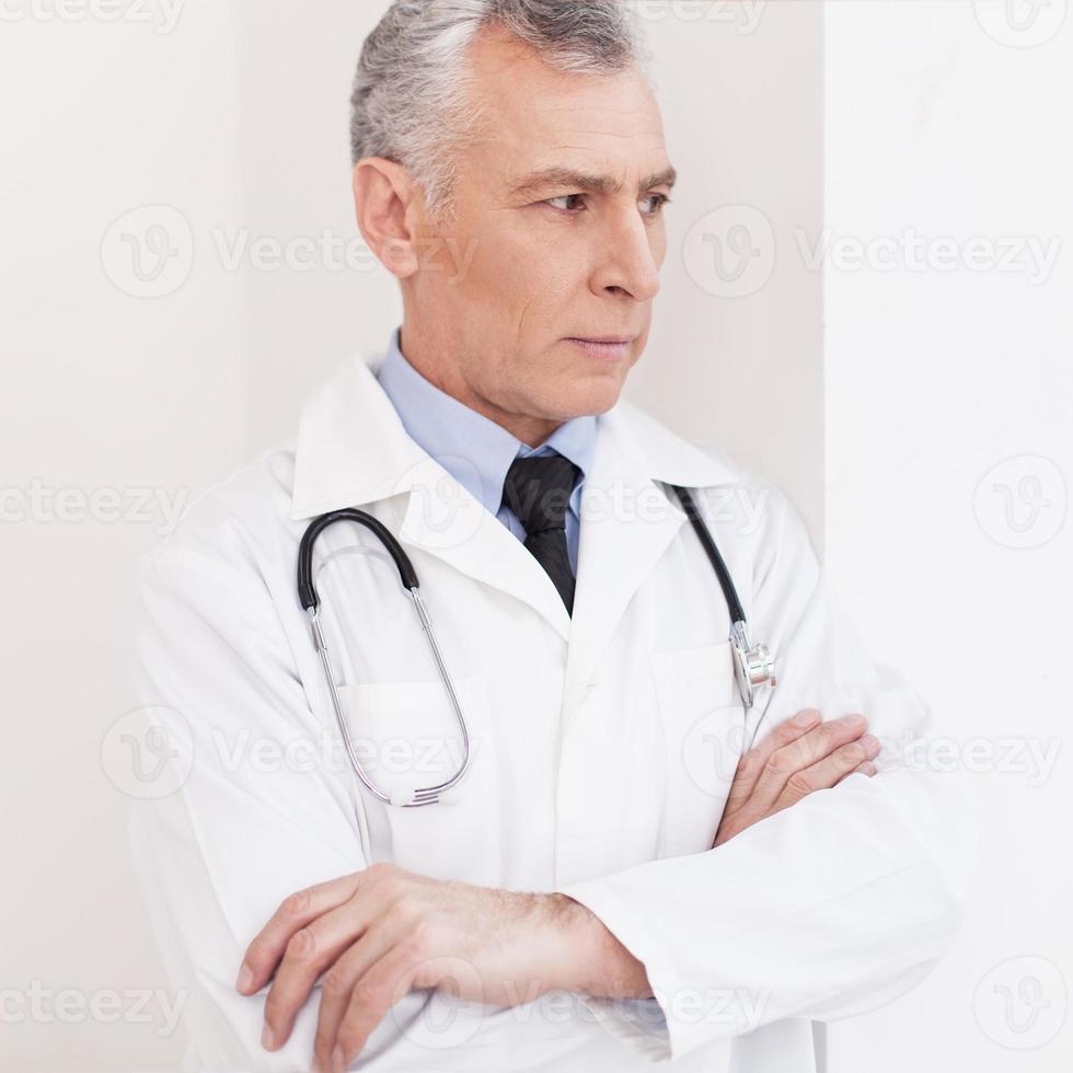 médico atencioso. médico sênior de cabelos grisalhos de uniforme olhando para longe e mantendo as mãos entrelaçadas foto