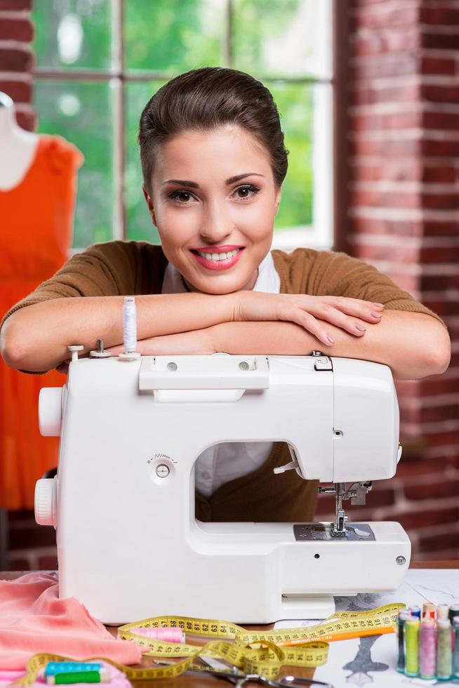 designer de moda confiante. bela estilista feminina inclinando-se para a máquina de costura e sorrindo enquanto está sentado em seu local de trabalho foto