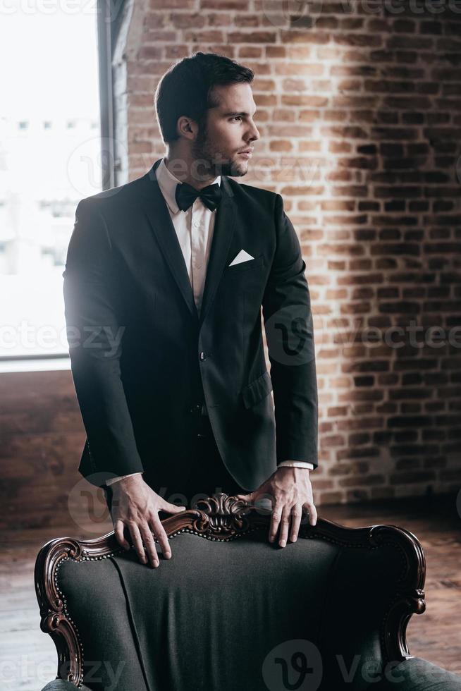 elegância e confiança. pensativo jovem bonito em traje formal e gravata borboleta, inclinando-se para a cadeira e desviar o olhar em pé no interior do loft foto