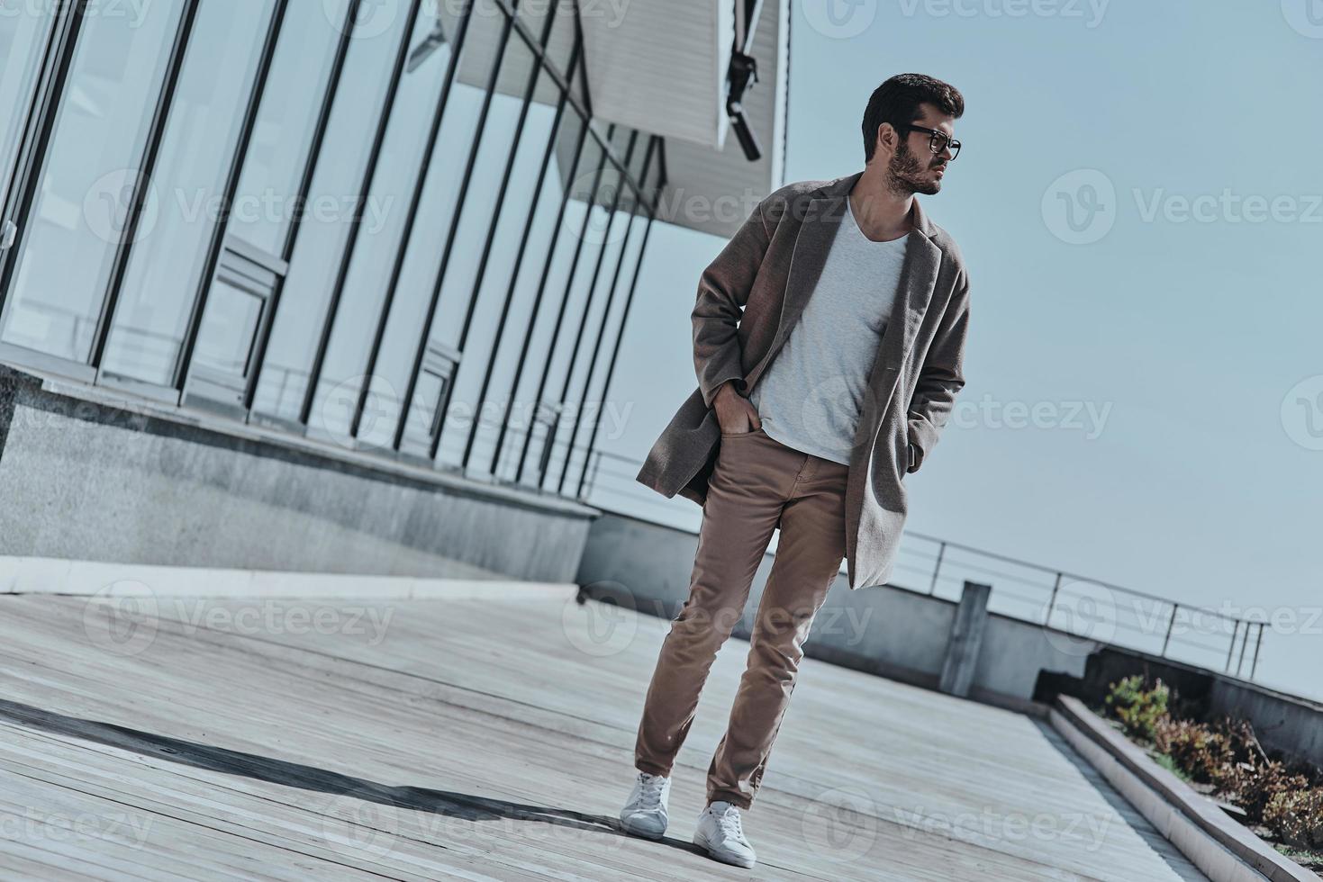 caminhada despreocupada. comprimento total de pensativo jovem moderno olhando para longe e mantendo as mãos nos bolsos enquanto caminha ao ar livre foto