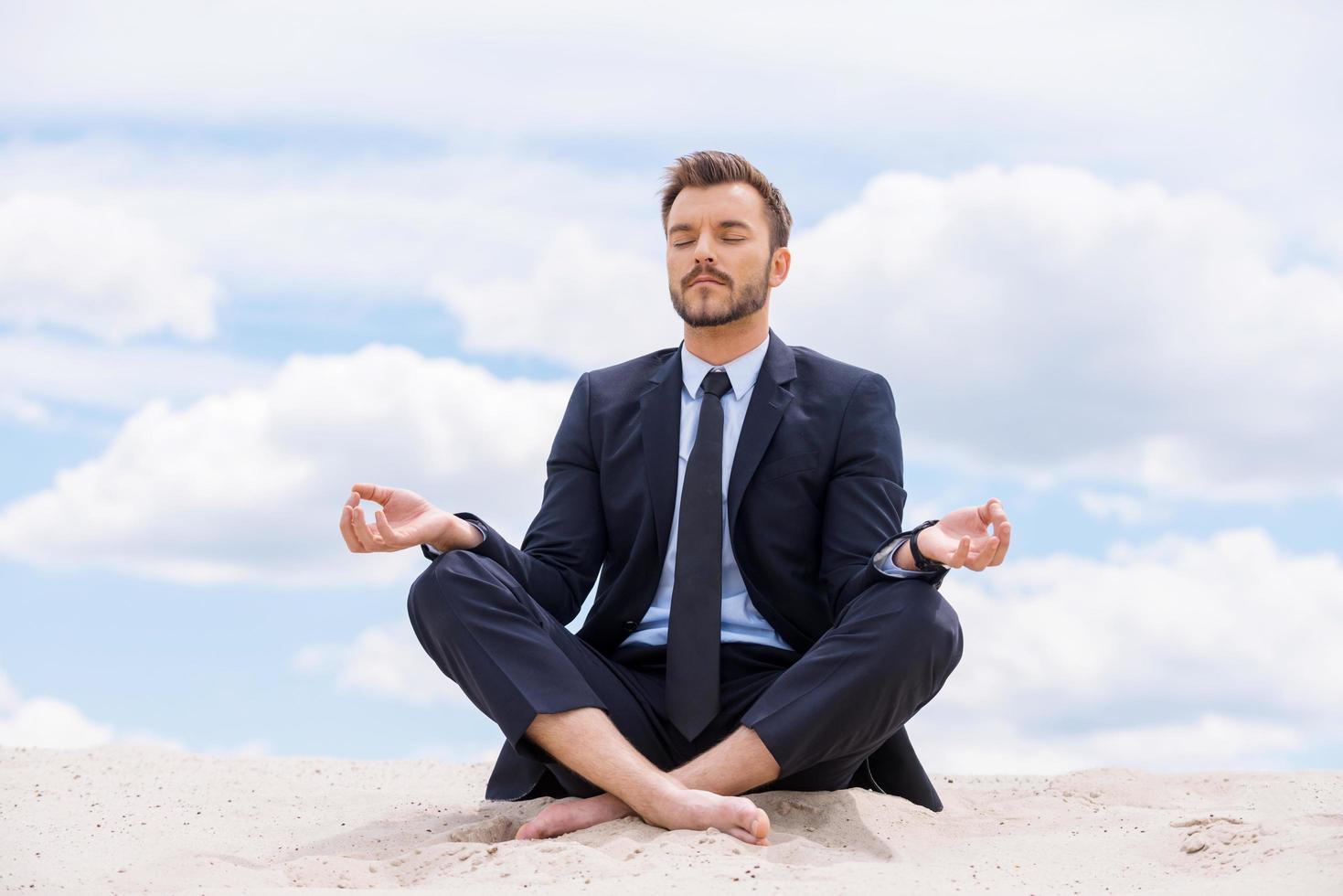 mantendo a calma dentro de sua alma. bonito jovem empresário meditando enquanto está sentado em posição de lótus na areia e contra o céu azul foto