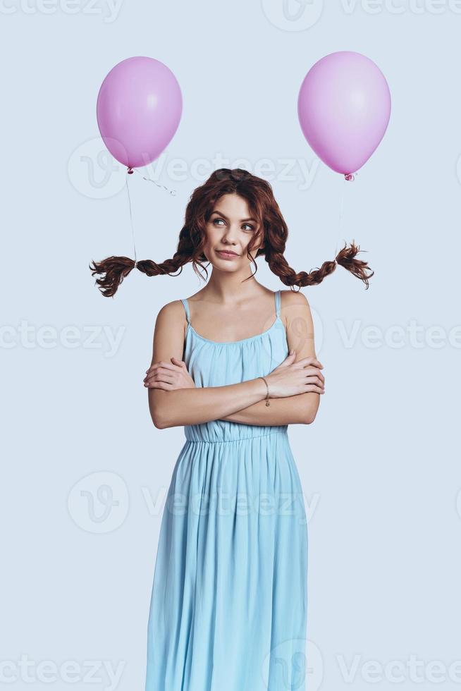 e se... tiro de estúdio de mulher jovem e bonita com balões amarrados às suas tranças sorrindo e olhando para a câmera em pé contra um fundo cinza foto