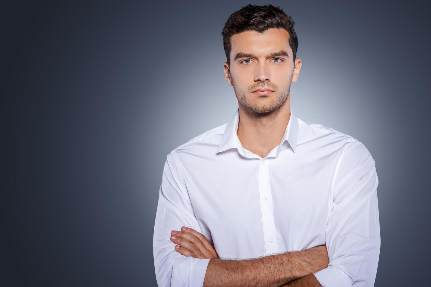 confiante bonito. jovem bonito na camisa branca, olhando para a câmera e mantendo os braços cruzados em pé contra um fundo cinza foto