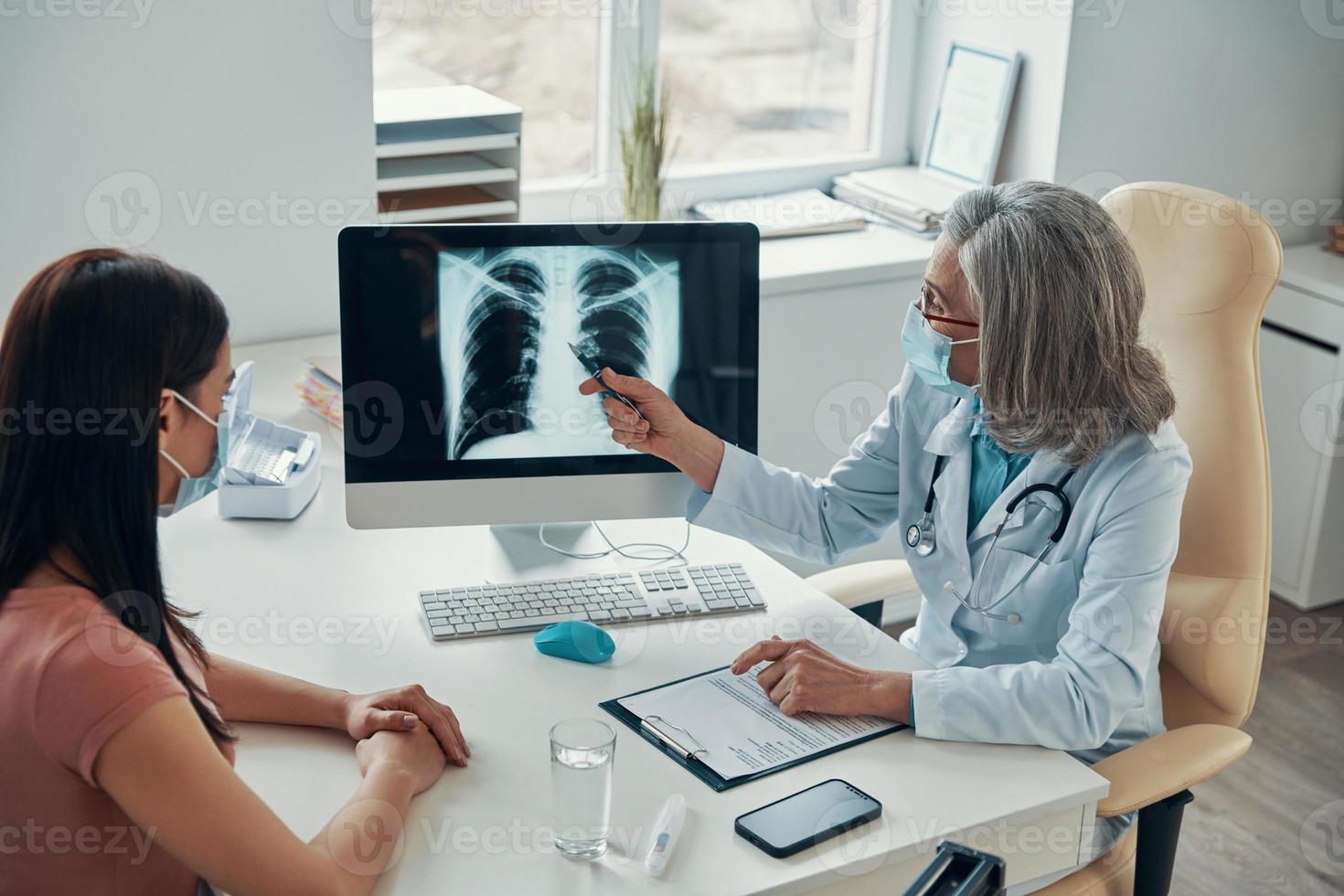 médica madura em jaleco branco mostrando para jovem seus pulmões escanear enquanto está sentado em seu escritório foto