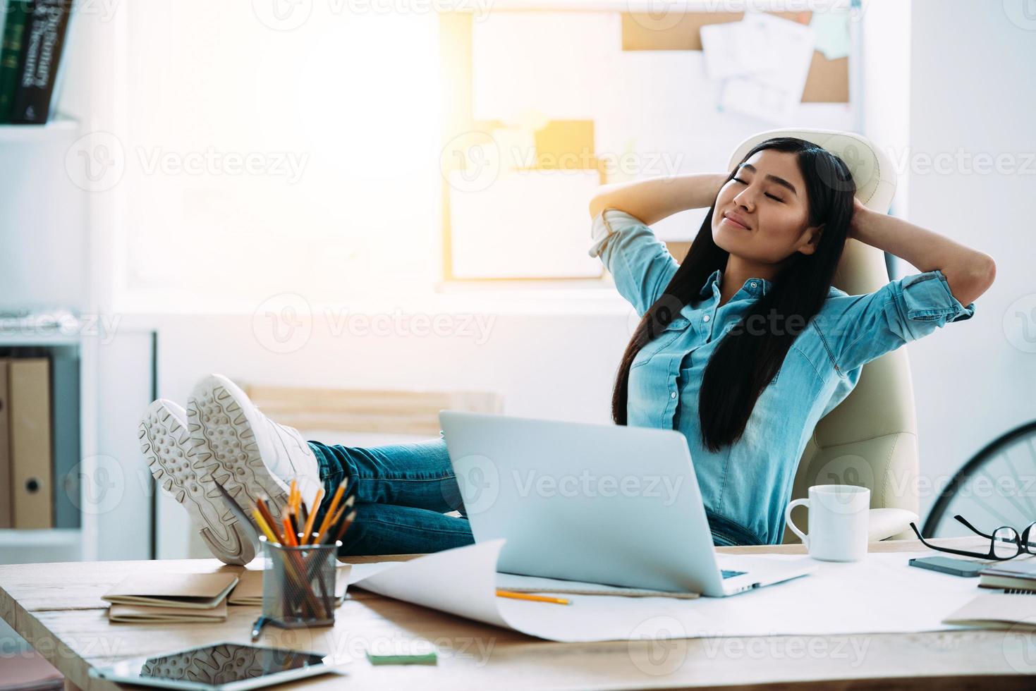 tendo um bom descanso. bela jovem asiática mantendo as mãos atrás da cabeça e parecendo relaxada enquanto está sentado em seu local de trabalho foto