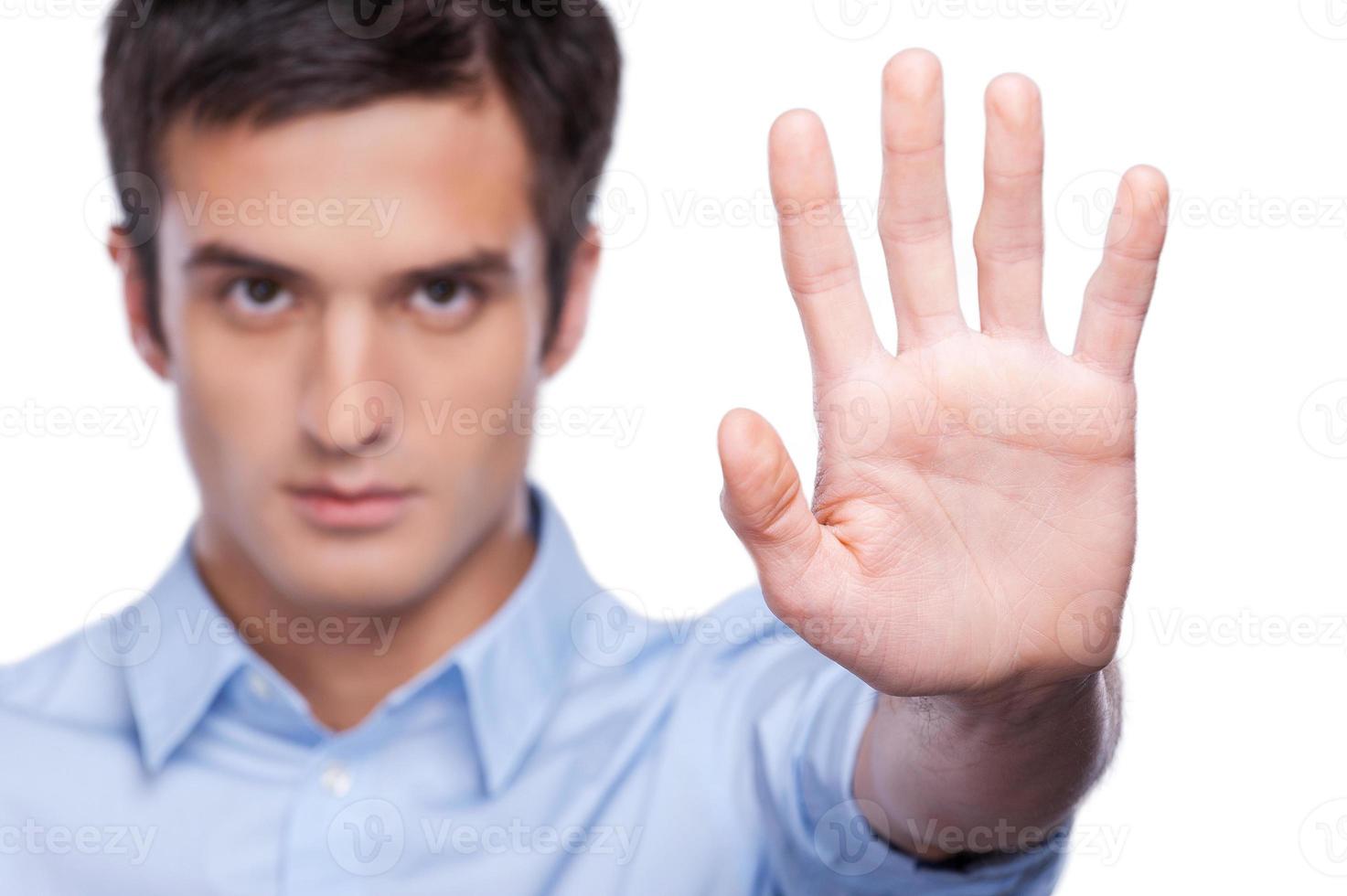 Pare o jovem sério na camisa azul, mostrando a palma da mão e olhando para a câmera em pé isolado no branco foto