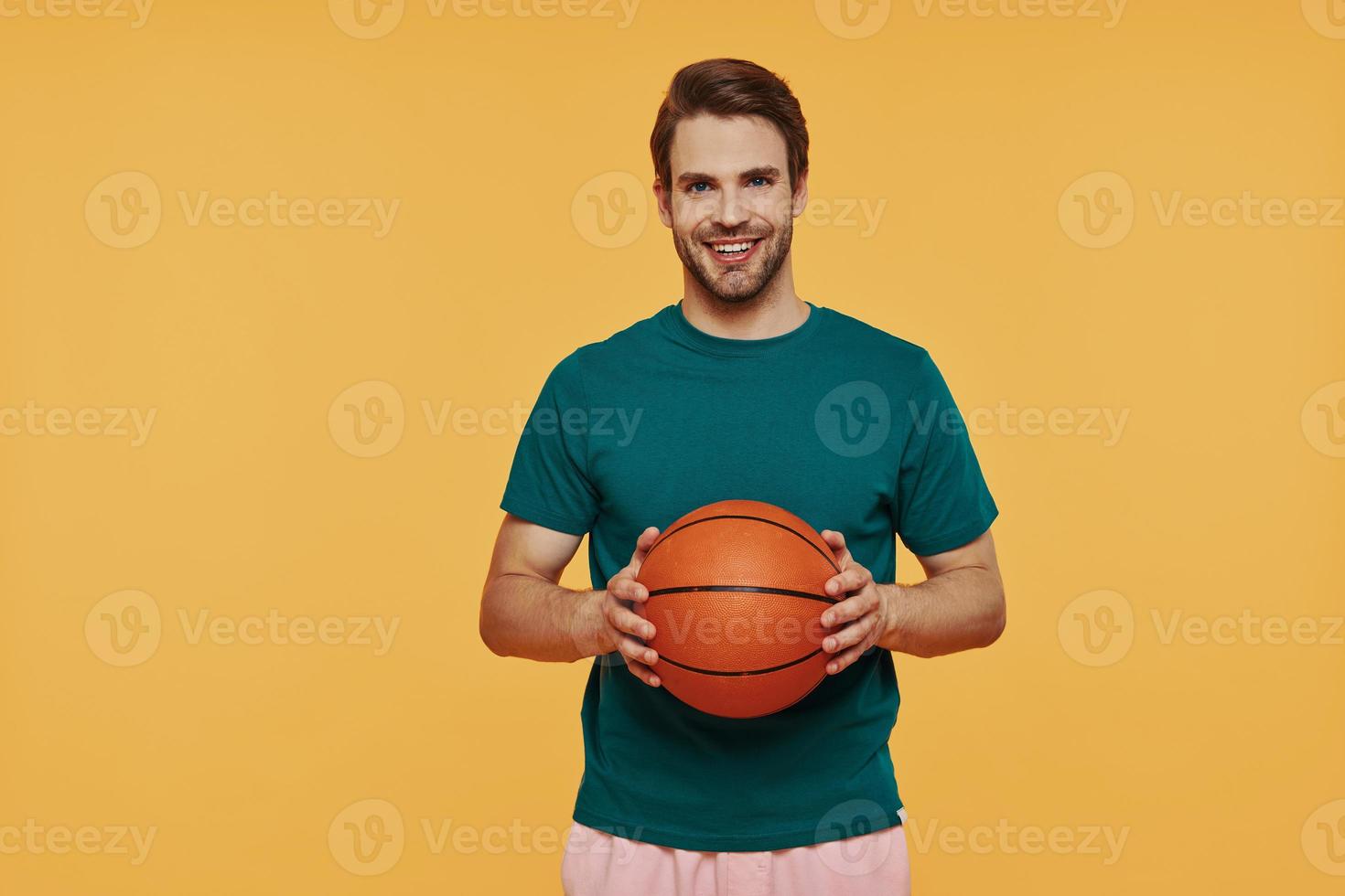 jovem bonito segurando uma bola de basquete e sorrindo em pé contra um fundo amarelo foto