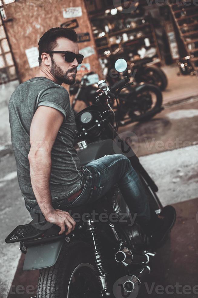 a liberdade está em suas veias. vista traseira do jovem confiante sentado em sua bicicleta e olhando por cima do ombro com garagem para motos ao fundo foto