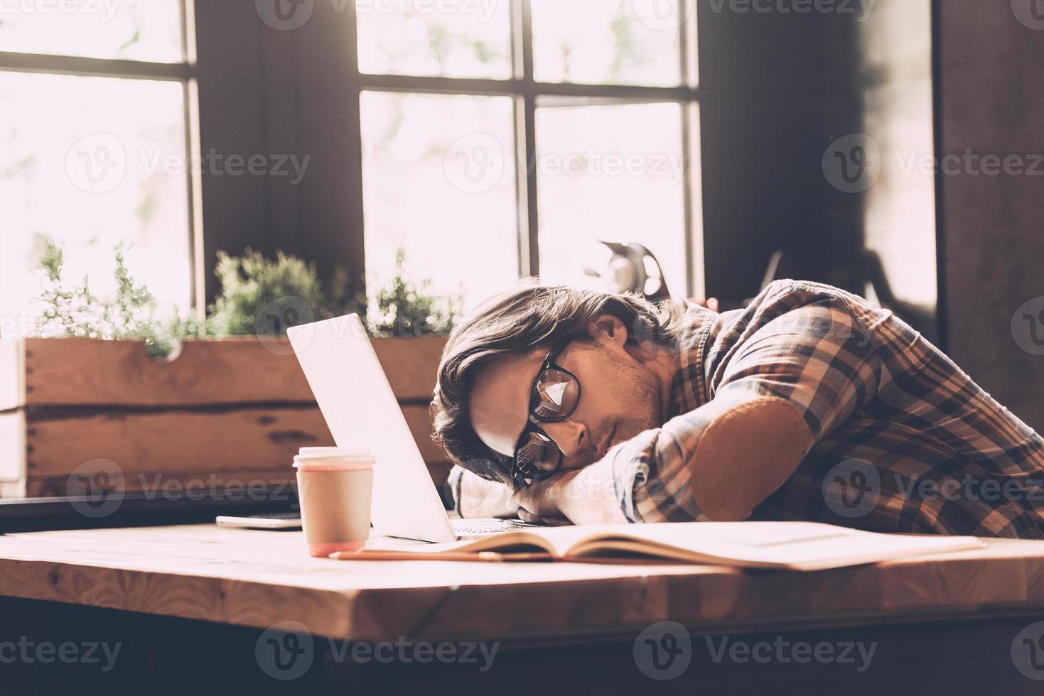 sentindo tão cansado. jovem em roupas casuais, inclinando a cabeça no laptop e dormindo enquanto está sentado em seu local de trabalho no escritório criativo foto