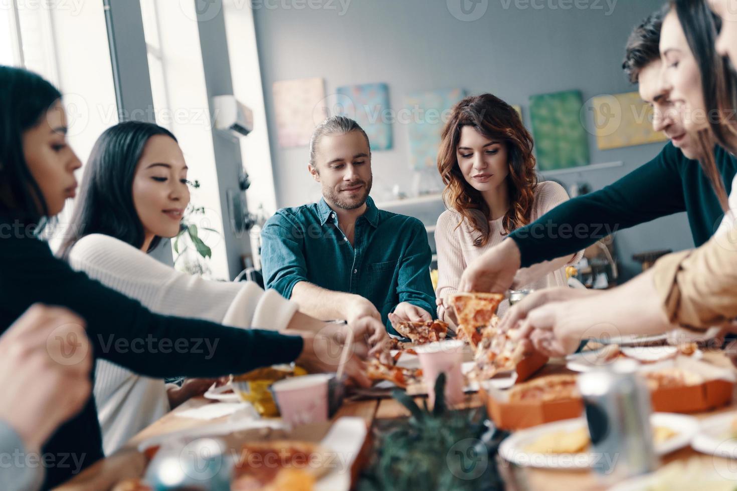 grupo de jovens em roupas casuais escolhendo pizza e sorrindo enquanto faz um jantar dentro de casa foto