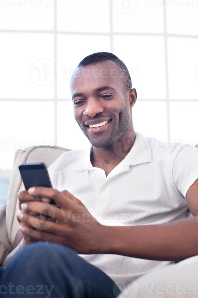 homem com telefone celular. alegre homem africano digitando algo no celular e sorrindo enquanto está sentado na cadeira foto