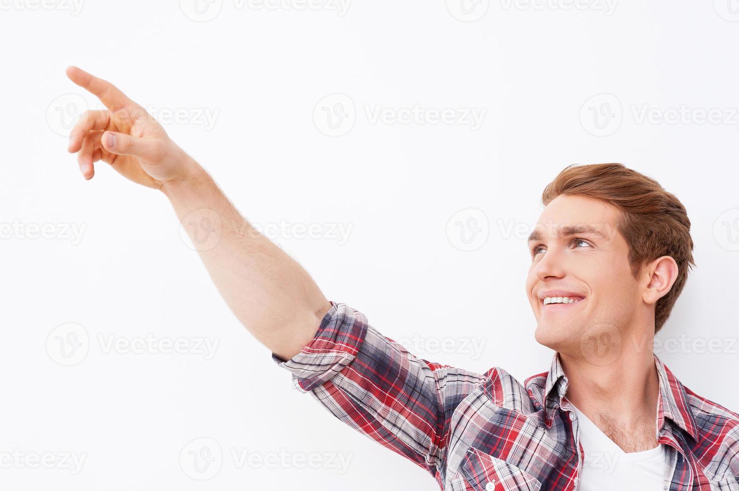 olhe ali jovem bonito apontando para longe e sorrindo em pé contra um fundo branco foto