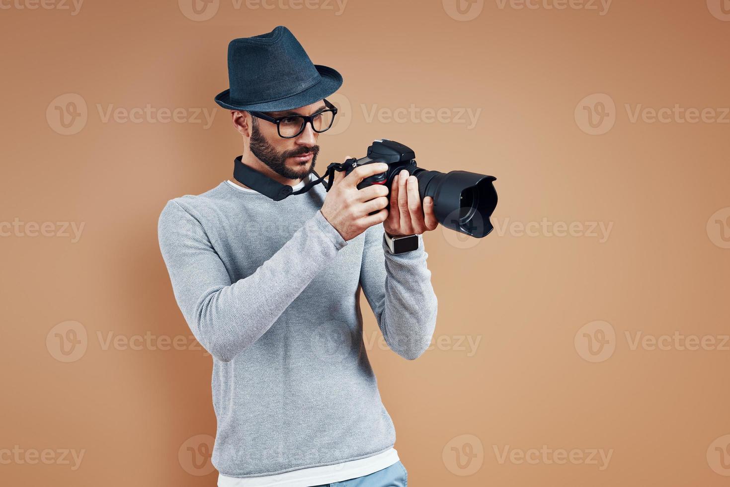 encantador jovem em roupas casuais, ajustando a câmera fotográfica em pé contra um fundo marrom foto