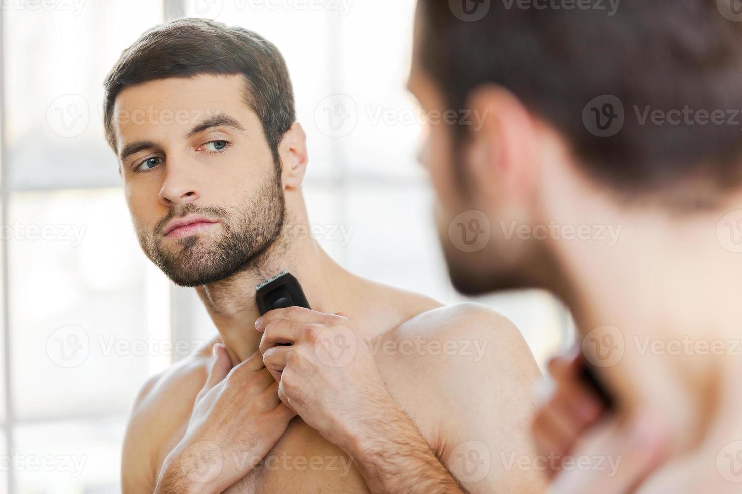 procedimento de barbear. vista traseira do jovem bonito barbear o rosto com barbeador elétrico em pé na frente do espelho foto