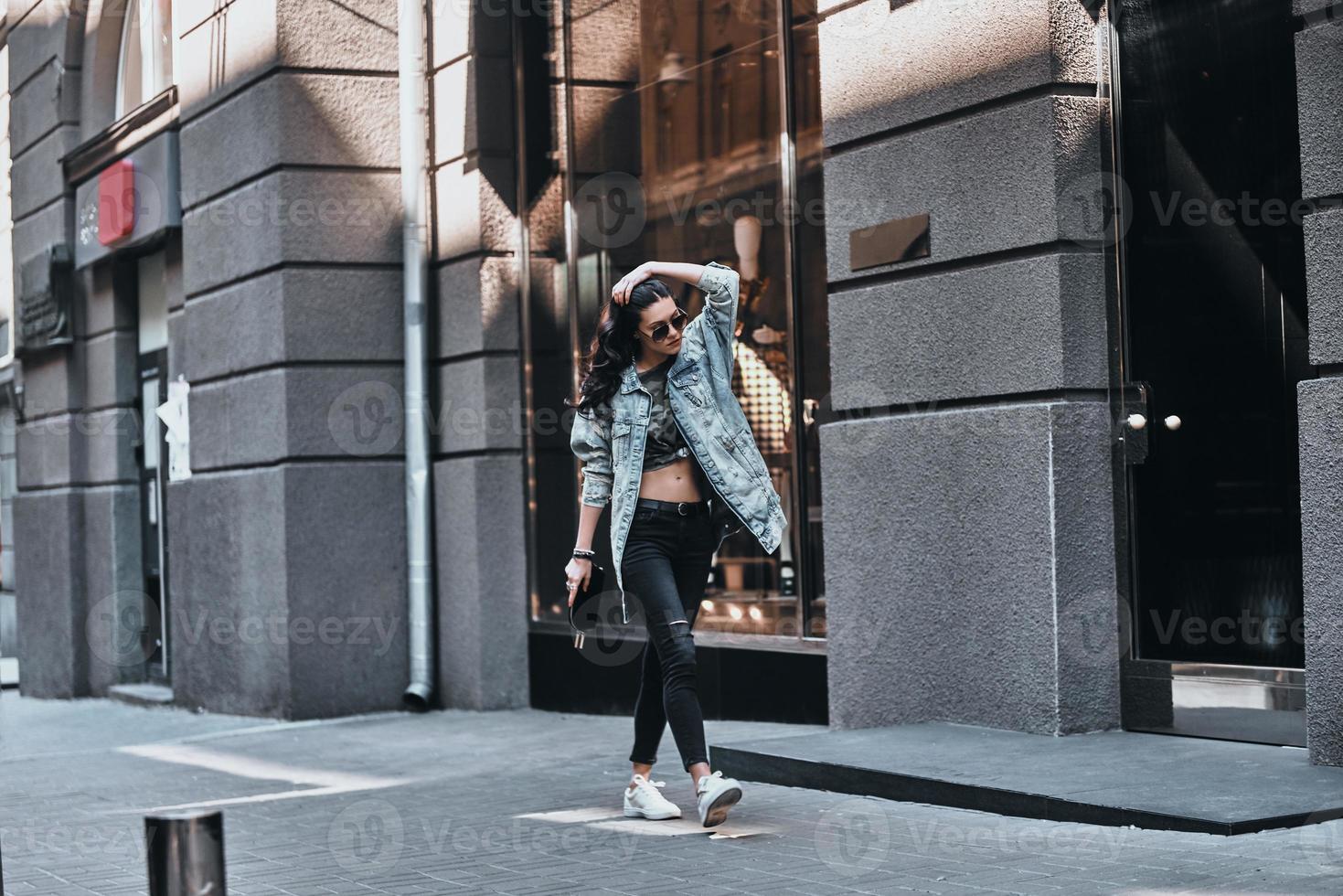 ótima aparência. comprimento total de mulher jovem e bonita na jaqueta jeans, mantendo a mão no cabelo enquanto caminhava pela rua foto