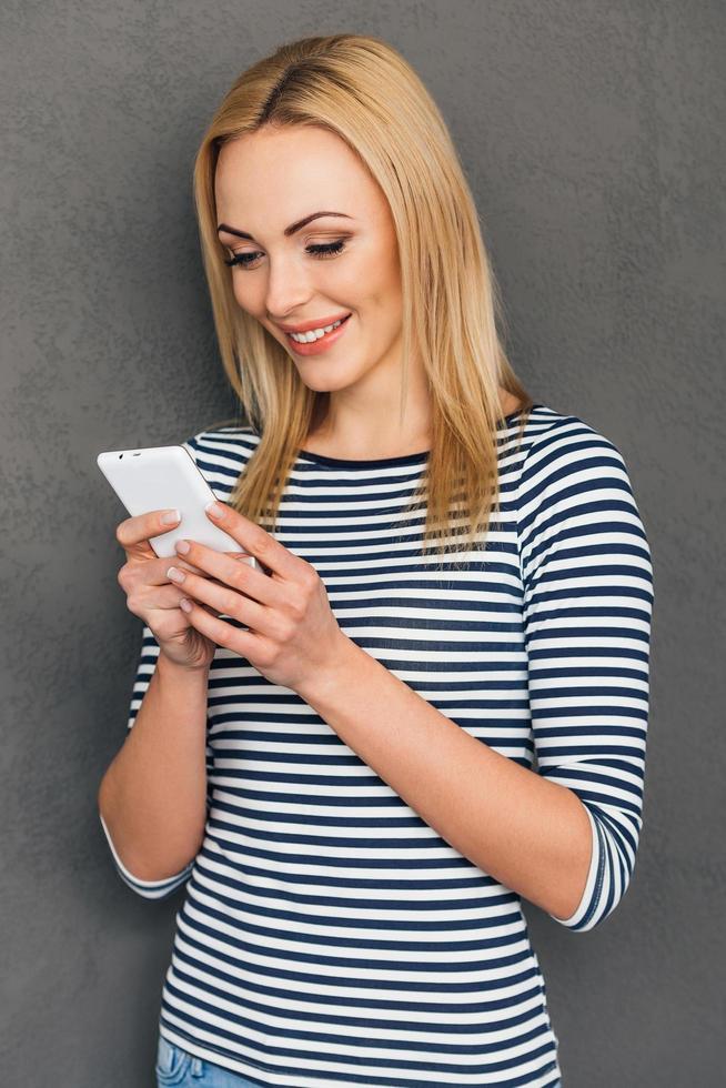 mensagem para amiga. bela jovem usando seu telefone inteligente e olhando para ele com um sorriso em pé contra um fundo cinza foto