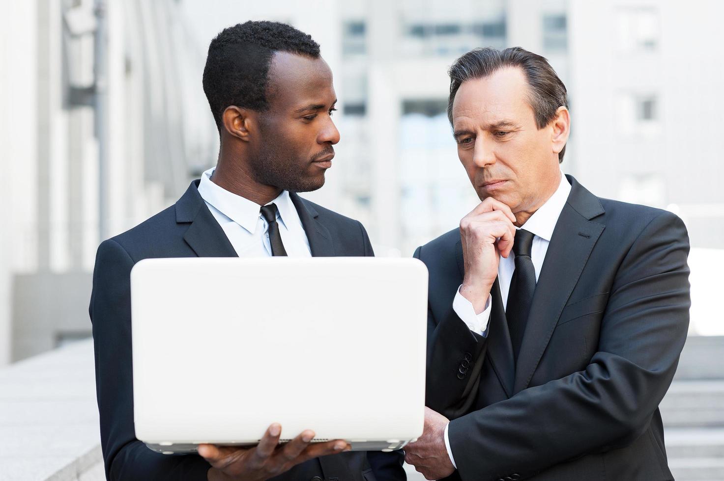 ele precisa de um conselho especializado. dois homens de negócios confiantes olhando para laptop enquanto um deles segurando a mão no queixo foto