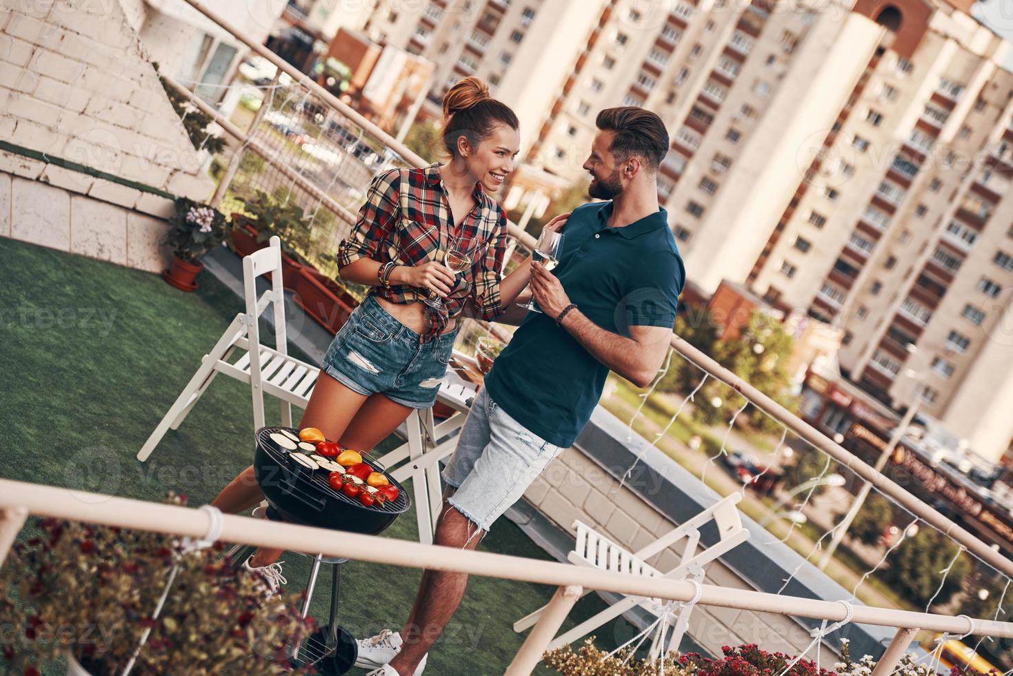 charmoso casal jovem em roupas casuais preparando churrasco e sorrindo em pé no pátio da cobertura ao ar livre foto