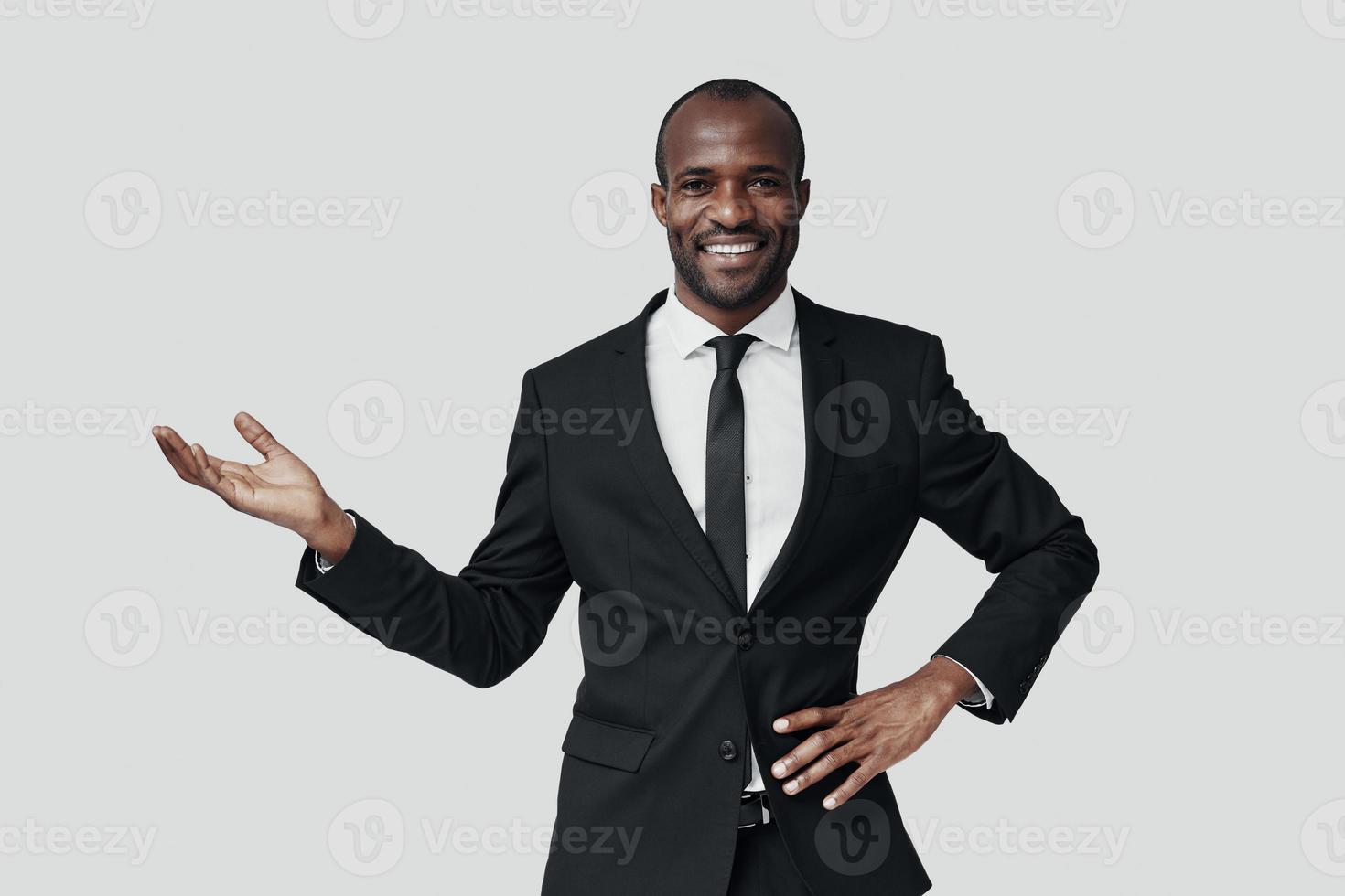 encantador jovem africano em trajes formais, apontando o espaço da cópia e sorrindo em pé contra um fundo cinza foto