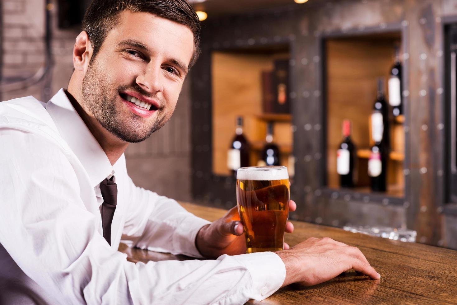 desfrutando de cerveja gelada e fresca. Vista lateral do jovem bonito de camisa e gravata segurando o copo com cerveja e sorrindo enquanto está sentado no balcão do bar foto