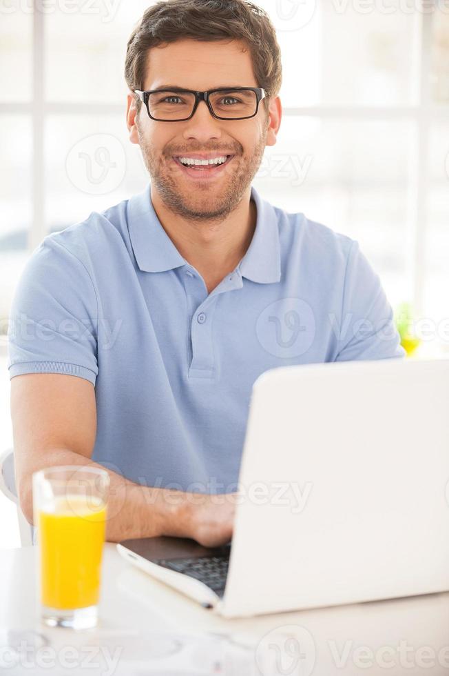 eu amo trabalhar em casa jovem bonito trabalhando no laptop e sorrindo enquanto está sentado à mesa com um copo de suco de laranja nele foto