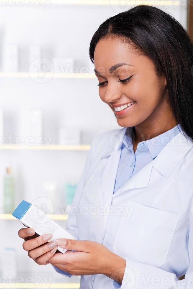 examinando novos medicamentos. bela jovem africana no jaleco segurando o recipiente com algum remédio e olhando para ele com um sorriso em pé na farmácia foto