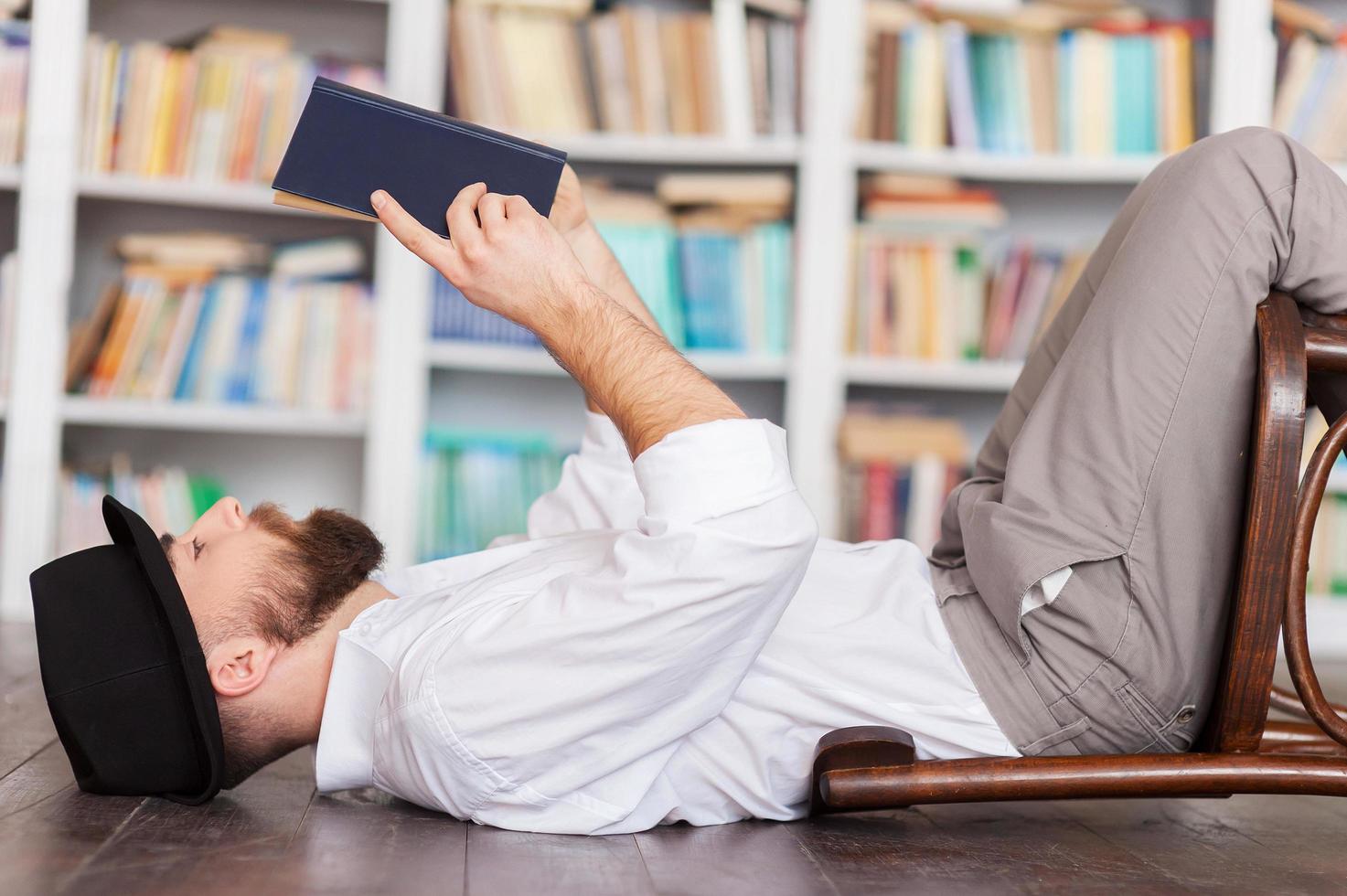 lindo leitor de livros. vista lateral do jovem pensativo de camisa e suspensórios deitado no chão e lendo um livro foto