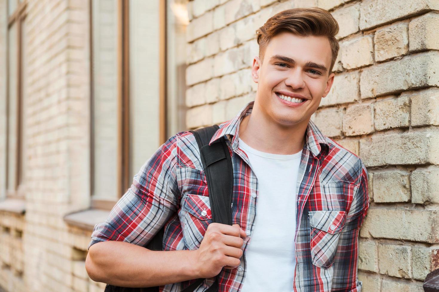 pronto para estudar. jovem bonito carregando mochila em um ombro e sorrindo enquanto se inclina para a parede de tijolos foto