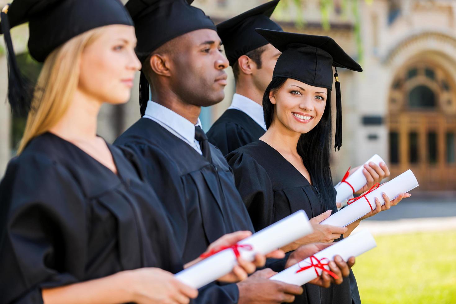 feliz graduado. quatro graduados em uma fila e segurando seus diplomas enquanto uma mulher olhando para a câmera e sorrindo foto