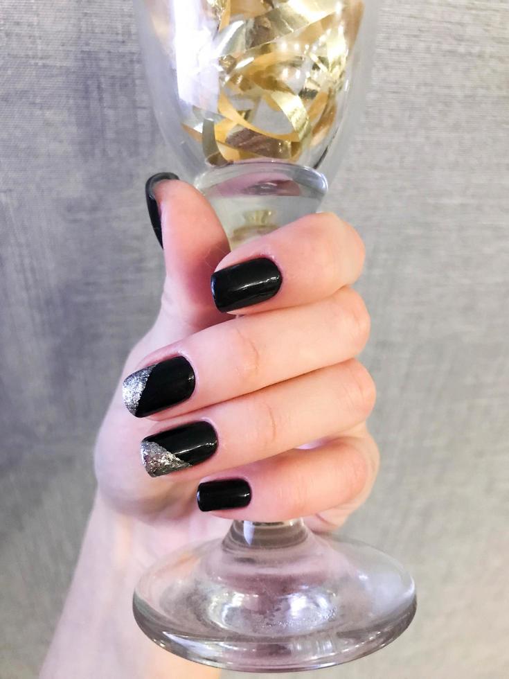 linda mão feminina com dedos finos e manicure preta com verniz gel com brilhos prateados nas unhas segurando um copo de vinho foto