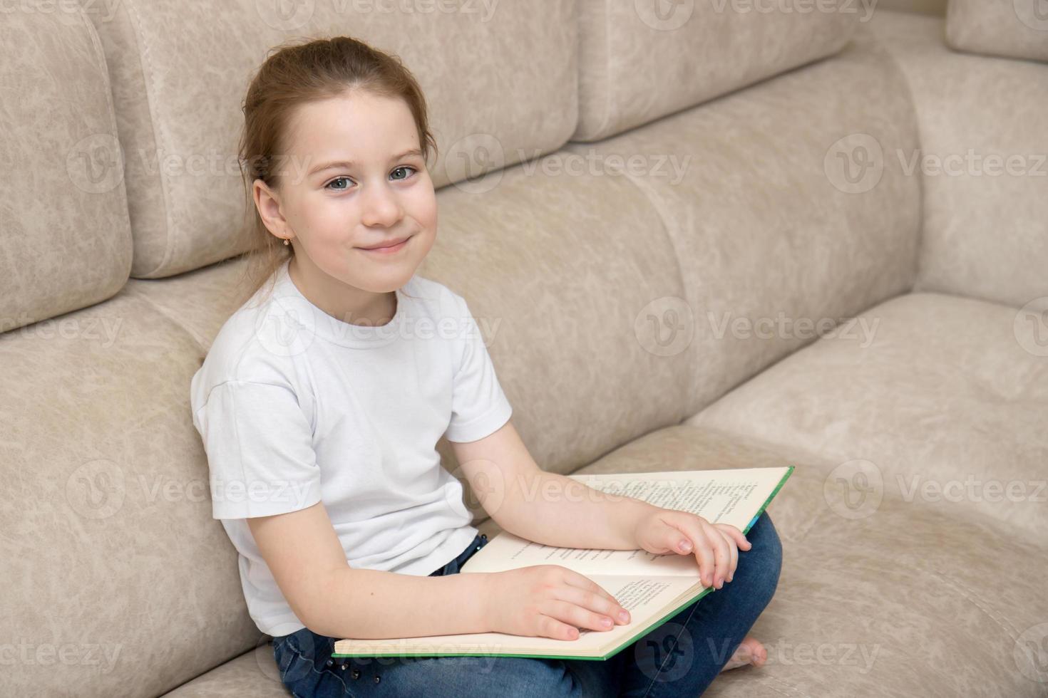 menina bonita senta-se em uma sala em um sofá em estilo turco segura um livro de joelhos e sorri, o conceito de aprendizagem, leitura, hobbies, volta às aulas foto